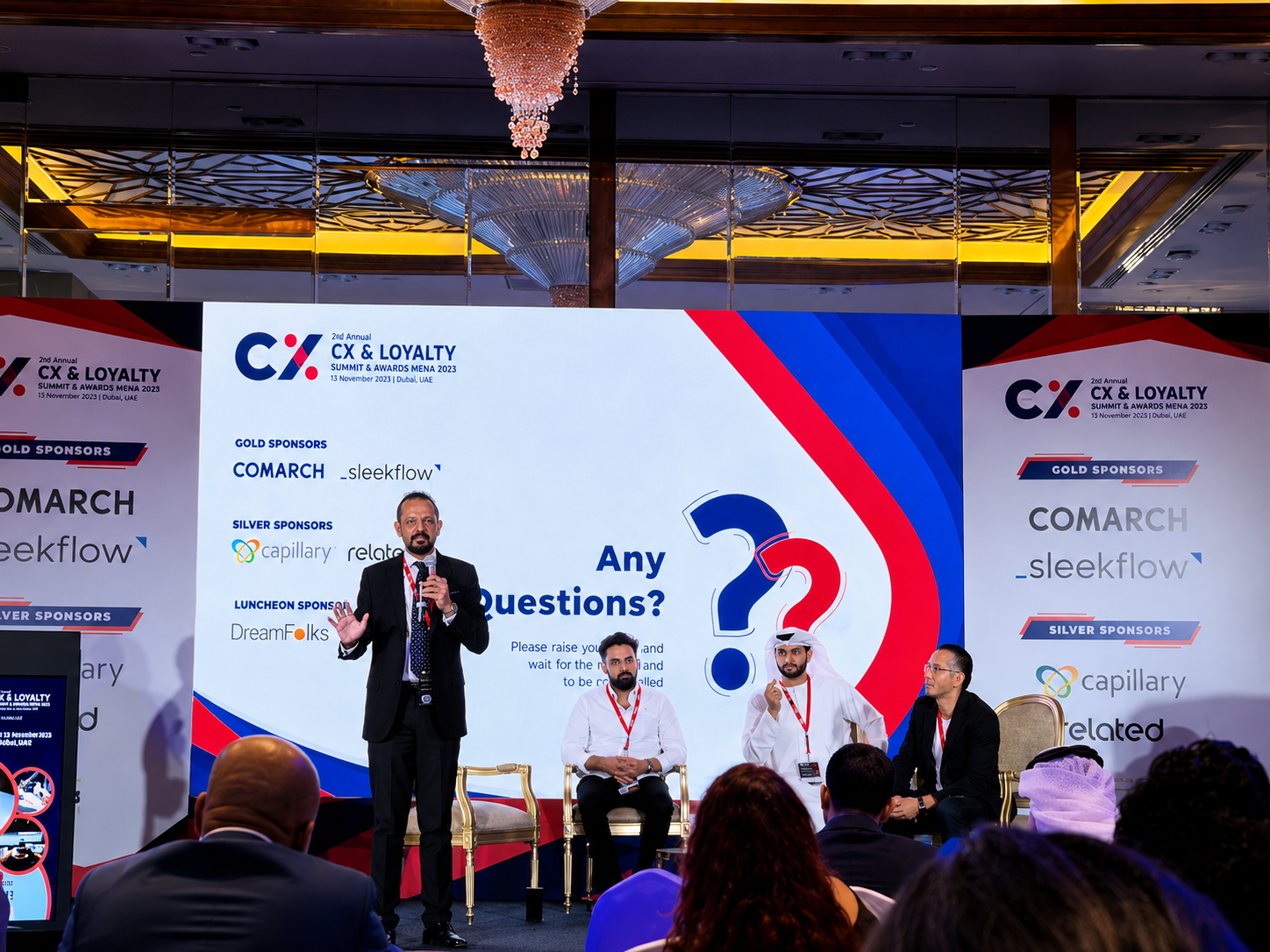 A speaker addressing the audience beside an Any Questions? slide on stage at a CX and Loyalty Summit