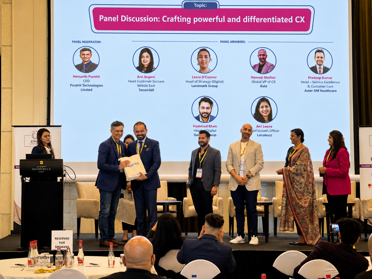 Two panellists exchanging a gift bag on stage in front of a customer experience panel discussion slide