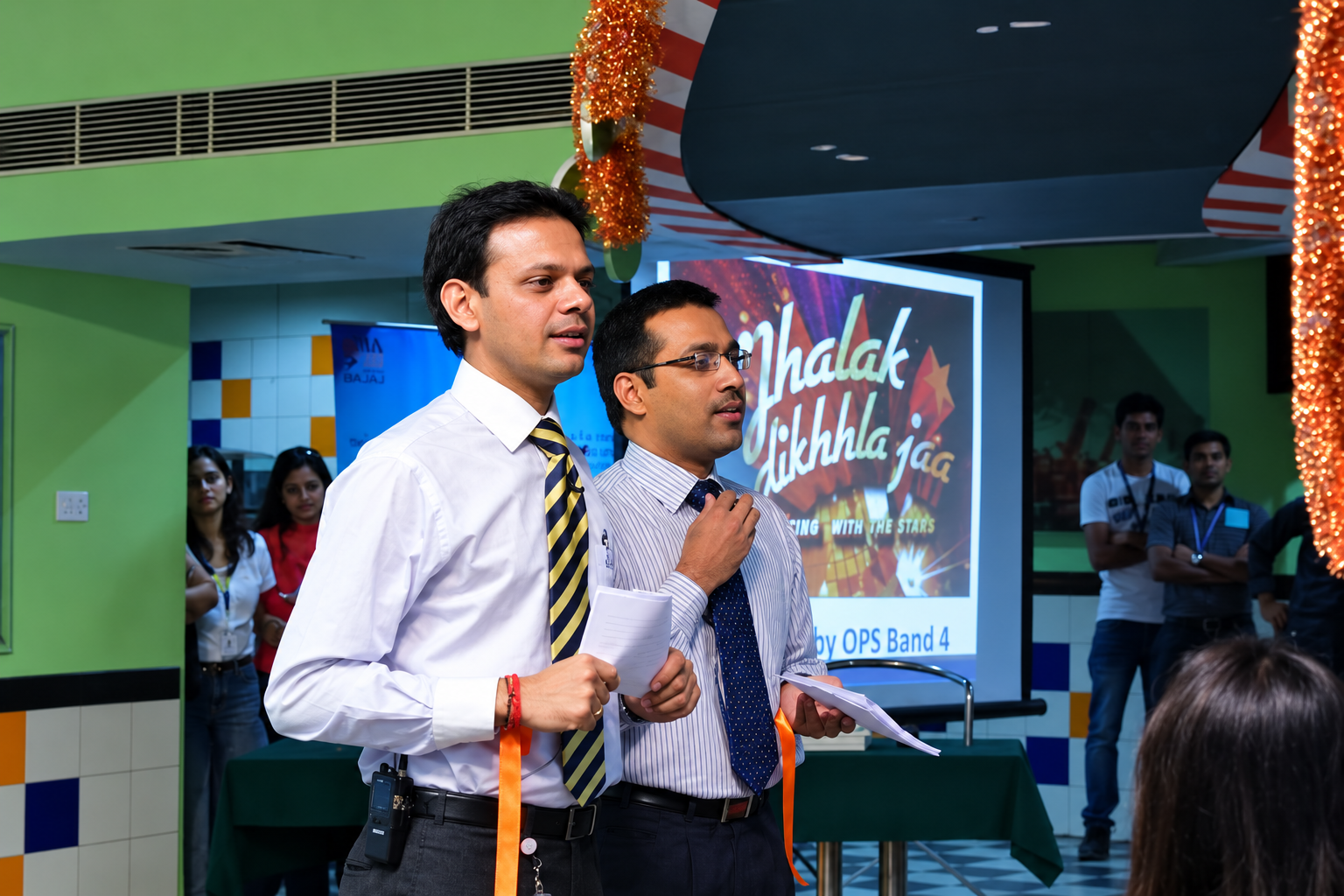 Two colleagues speaking into a microphone in front of a Jhalak Dikhhla Jaa dance competition screen at an office event