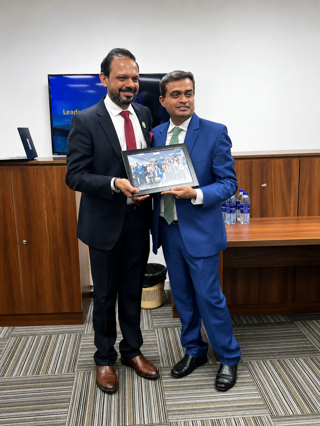 Two colleagues holding a framed team photograph in front of a Leader's Day screen