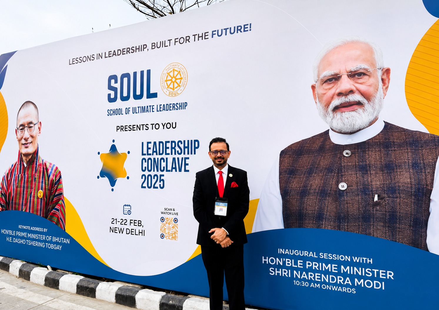 A man in a suit standing in front of a School of Ultimate Leadership Conclave 2025 banner featuring Indian Prime Minister Narendra Modi and Bhutan's Prime Minister Dasho Tshering Tobgay